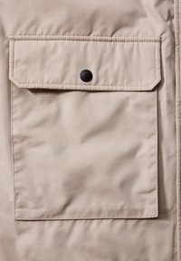 Poche en tissu beige avec rabat fixé par un seul bouton-pression noir sur une surface de vêtement matelassée.