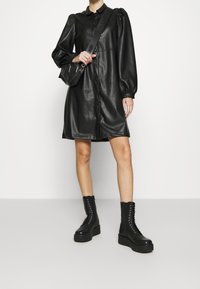 Robe boutonnée en similicuir noir avec manches bouffantes et coupe décontractée. Associée à des bottes noires épaisses à lacets pour un contraste marqué.