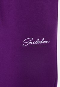 Fialová látková mikina s bílým vyšívaným logem "Smélodox" a zeleným akcentem. Hladký povrch, jednoduchý design, žádné další vzory.