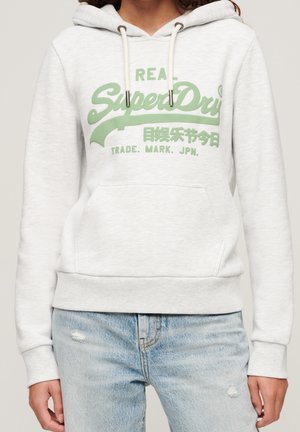White hooded sweatshirt with green "SuperDry" logo and light blue ripped jeans on a person cropped at the neck.