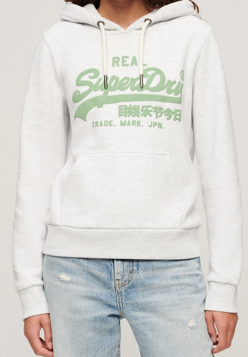 Sweatshirt à capuche blanc avec logo "SuperDry" vert et jean déchiré bleu clair sur une personne cadrée au niveau du cou.
