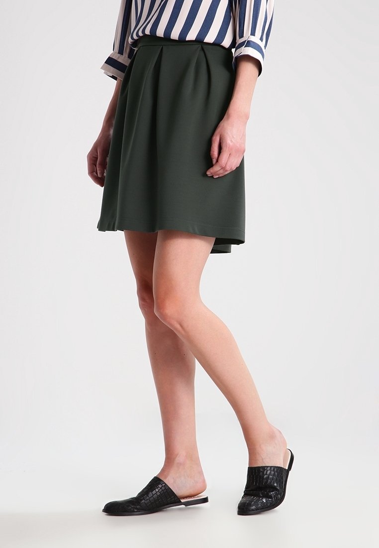 Femme portant une jupe plissée vert foncé, un chemisier à manches longues rayé et des chaussures noires sans lacets, debout, jambes croisées sur un fond blanc.
