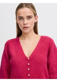 Fukšijas krāsas adīts džemperis ar V-veida kakla izgriezumu un garām piedurknēm, kas rotāts ar pieciem baltiem pērļu līdzīgiem pogām gar priekšpusi un ar rakstainu faktūru.