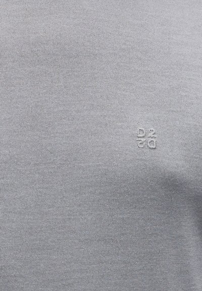 Chemise en tissu tricoté gris avec un logo discret en haut à gauche. La texture semble lisse avec un design léger.