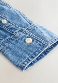 Lyseblå denimskjorte med manchetter, der har to hvide knapper, synlig syning og en let slidset kant for tekstur og detaljer.