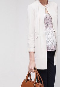 Jasnobeżowa kurtka z okrągłym dekoltem i rękawami do łokcia, noszona na bluzce w kwiatowy wzór. Brązowa torebka trzymana w dłoni.