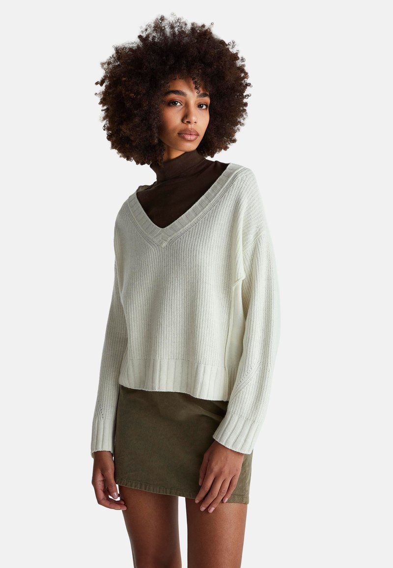 V-Ausschnitt, gerippter Strickpullover in Off-White, kombiniert mit einem olivgrünen Cord-Minirock. Darüber liegt ein brauner Rollkragenpullover.