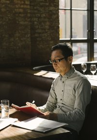 Un uomo in una camicia grigia chiara scrive su un quaderno rosso a un tavolo di legno con un bicchiere d'acqua e dei fogli nelle vicinanze, mentre la luce del sole filtra dentro.