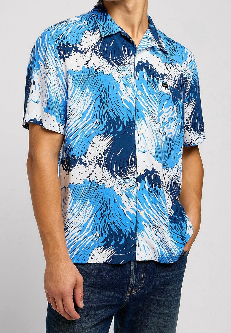 Muž oblečený v košili s krátkým rukávem s modro-bílým vlnkovým vzorem, doplněný tmavě modrými džíny.