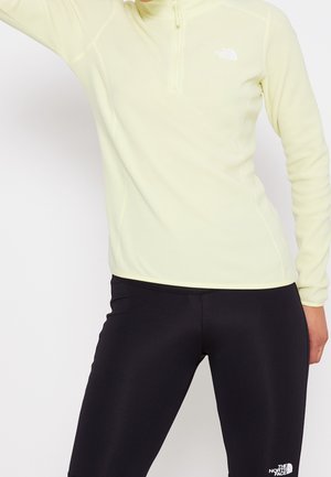 Persona con una camiseta de manga larga color amarillo pálido con cremallera en el cuello y leggings negros con logos de The North Face, posando con un brazo levantado.