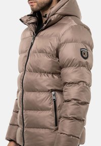 Gepolsterte Winterjacke in einem hellbraunen Farbton, mit einer Kapuze, einem Reißverschluss vorne und seitlichen Taschen. Enthält ein Logo-Patch am Ärmel.