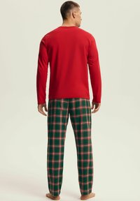 Mężczyzna stojący boso, ubrany w czerwoną bluzę z długim rękawem i zielono-czerwono-białe, kraciasto wzorzyste spodnie do piżamy, zwrócony tyłem na jednolitym tle.