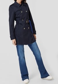 Námořnický modrý trenčkot s dvojitými knoflíky, širokými klopami, páskem a dvěma předními kapsami; kombinovaný s rozšířenými modrými džíny a bílými teniskami.