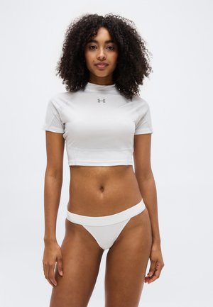 Vrouw met krullend haar, gekleed in een wit cropped Under Armour T-shirt en witte bikinibroekjes, tegen een effen witte achtergrond.