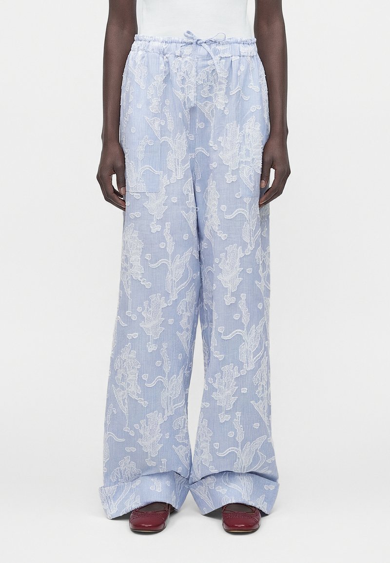 Φαρδιά παντελόνια ανοιχτού μπλε χρώματος με λευκή κεντητή λουλουδένια διακόσμηση, ελαστική μέση με κορδόνι, φορεμένα με σκοτεινά κόκκινα επίπεδα παπούτσια.