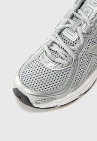 Zapatilla deportiva gris con parte superior de malla transpirable, puntera reforzada, detalles reflectantes y suela acolchada. Presenta cordones texturizados.
