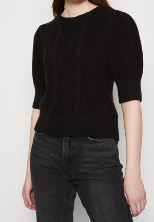 Mujer con un suéter negro de punto trenzado, mangas abullonadas y dobladillo acanalado, combinado con jeans negros de talle alto.