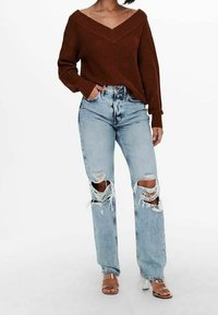 Pull marron en maille avec col en V, associé à un jean bleu clair déchiré avec de grandes déchirures aux genoux et des sandales à talons transparents.