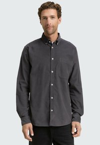 Camisa de manga larga y botones en corduroy gris oscuro con un solo bolsillo en el pecho y botones blancos. Textura suave y diseño de cuello estándar.
