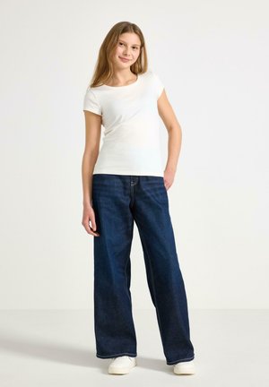 Ung kvinde står med den ene hånd i lommen, iført hvid kortærmet skjorte, mørkeblå jeans med vide ben og hvide sneakers på ensfarvet baggrund.