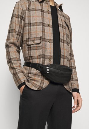 Hombre con camisa de cuadros beige y naranja abotonada, pantalones negros y riñonera negra con cremallera, de pie con las manos en los bolsillos.