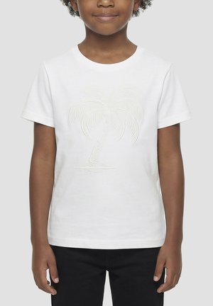 Niño con una camiseta blanca con un diseño texturizado de palmera, de pie frente a un fondo gris claro liso.