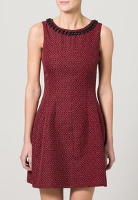 Robe sans manches rouge avec un motif noir complexe et un col orné de perles. Corsage ajusté, jupe évasée, tissu texturé.