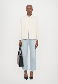 Giacca button-up beige chiaro, jeans a gamba larga blu chiaro con orlo sfrangiato, borsa nera con borchie e décolleté nere a punta.