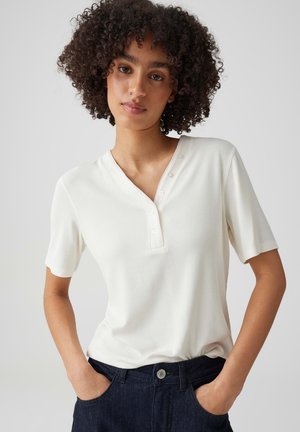Junge Person mit lockigem Haar trägt ein weißes Henley-Shirt mit kurzen Ärmeln und Knöpfen sowie dunkle Jeans, Hände in den Taschen.