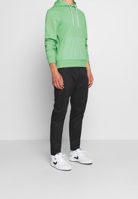 Persona che indossa un hoodie verde chiaro, pantaloni neri e scarpe da ginnastica bianche, in piedi contro uno sfondo grigio semplice.
