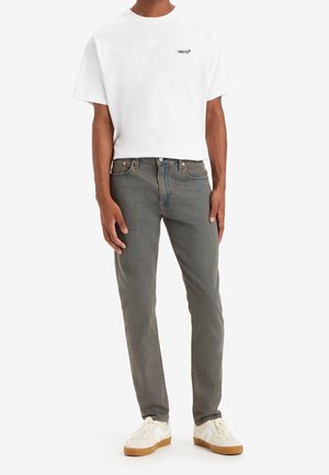 Mann trägt weißes Levi's T-Shirt, graue Jeans und weiße Sneakers mit Gummisohlen, steht vor weißem Hintergrund.