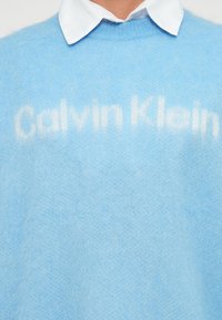 Világoskék kötött pulóver, puha anyagból készült, kerek nyakkivágással és a "Calvin Klein" logóval, egy kicsit világosabb árnyalatban.