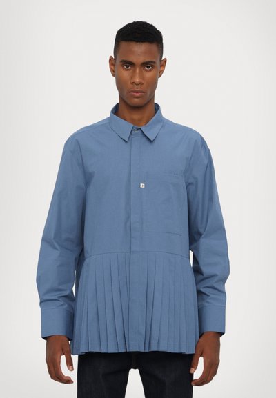Chemise bleue à manches longues avec col, présentant une partie inférieure plissée et une poche poitrine, fabriquée dans un tissu lisse.