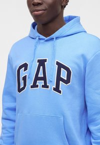 Jeune personne portant un sweat à capuche bleu clair avec des lettres bleu marine et blanches formant le mot GAP sur la poitrine, souriant légèrement.