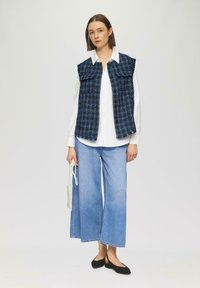 Blau- und schwarz kariertes Weste über einem weißen Hemd, kombiniert mit hellblauen weit geschnittenen Jeans und schwarzen Ballerinas. Enthält eine kleine weiße geflochtene Tasche.