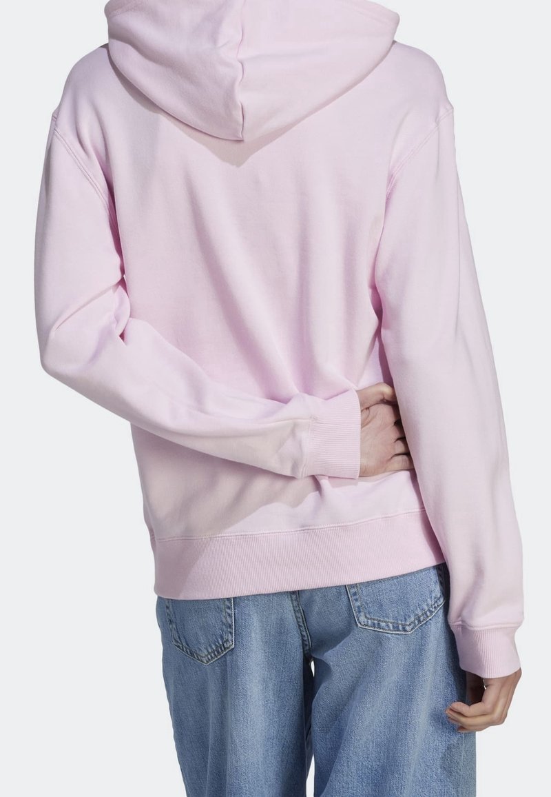 Rosa huvtröja med avslappnad passform, ribbade muddar och nederkant. Tyget verkar mjukt med minimala sysmån. Bärs med blå jeans.