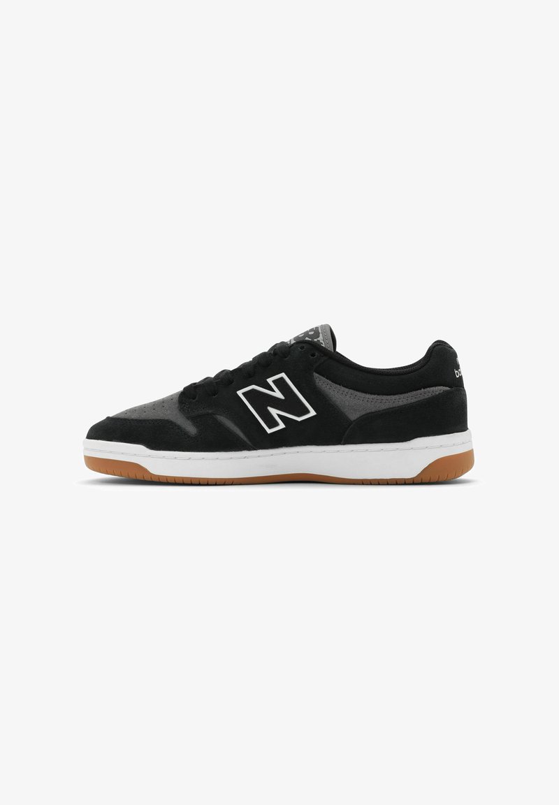 Zapatillas de deporte de ante negro y malla con acentos blancos, que cuentan con una suela de goma y un prominente logo 'N' en el lado. Diseño de perfil bajo.