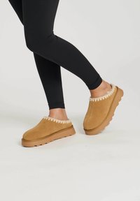 Sabots en daim marron avec un bord en fausse fourrure couleur crème, semelles rembourrées et un design à bout arrondi. Portés avec des leggings noirs.
