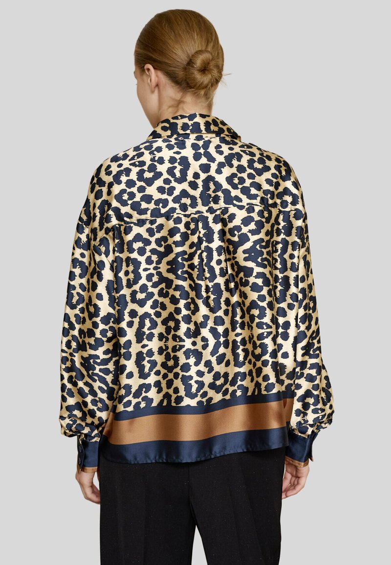 Kvinde med hår i en bun, iført en løstsiddende leopardmønstret bluse med marineblå og brun kant, vist fra ryggen mod en grå baggrund.