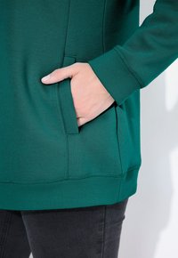 Felpa verde con una texture liscia, dotata di tasca laterale e polsini a costine. Anche l'orlo inferiore è a costine per una maggiore struttura.