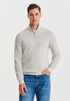 Jasnoszary sweter z półgolfem na zamek i teksturowanym wzorem dzianiny, zestawiony z niebieskimi dżinsami. Prosty, nowoczesny design.