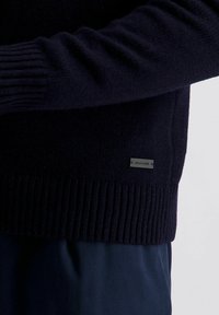 Dunkelblaues, geripptes Strickpullover mit Ärmel und Saum, versehen mit einem kleinen rechteckigen "Pierre Cardin" Metall-Logo-Tag an der Seite, über einer dunkelblauen Hose.