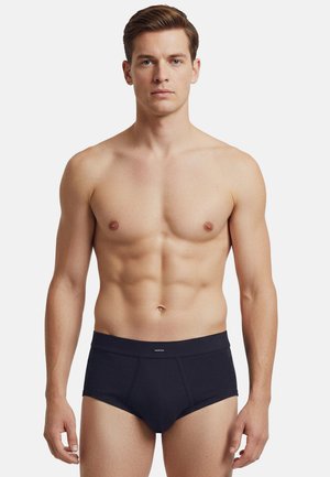 Veltrænet ung mand med kort brunt hår iført sorte underbukser, stående mod en ensfarvet hvid baggrund, vendt fremad med neutralt ansigtsudtryk.