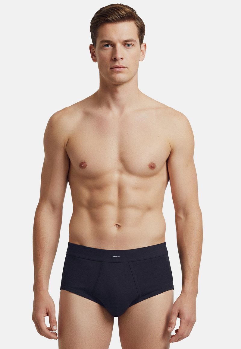 Giovane uomo in forma con capelli castani corti, indossa slip neri, in piedi davanti a uno sfondo bianco semplice, rivolto in avanti con espressione neutra.