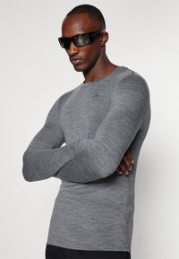 Camisola atlética cinza de mangas longas com um design ajustado, gola redonda e detalhe do logótipo no peito. O tecido parece suave e ligeiramente texturizado.