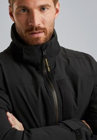 Giacca bomber nera con un alto collo, chiusura con zip, tessuto testurizzato, branding sottile e silhouette aderente.
