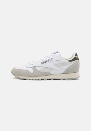 Bílá a světle šedá teniska Reebok s semišovými detaily, zavazováním na šněrování a logem na boku a patě, zobrazena z boku na jednobarevném pozadí.