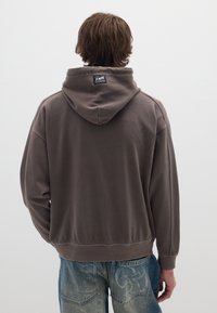 Felpa marrone con vestibilità rilassata, cappuccio e etichetta con marchio sul retro, abbinata a jeans azzurri chiari con motivi sensibili.