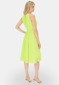 Sukienka do kolan, bez rękawów, wykonana z jasnej limonkowej chiflony, z dopasowaną talią i falbaniastym dołem. Przezroczyste sandały na obcasie dopełniają wizerunek.