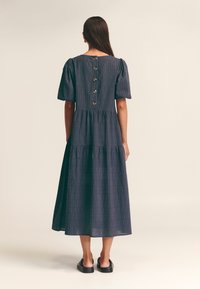 Donna che indossa un vestito midi blu navy con maniche a palloncino, linee verticali a pois e grandi bottoni lungo la schiena, abbinato a sandali neri senza chiusura.